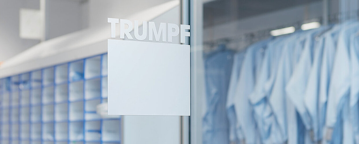 Innenansicht eines industriellen Raums mit blauen Regalen und hängenden blauen Arbeitskleidungen. An der Glastür steht der Schriftzug “TRUMPF”.