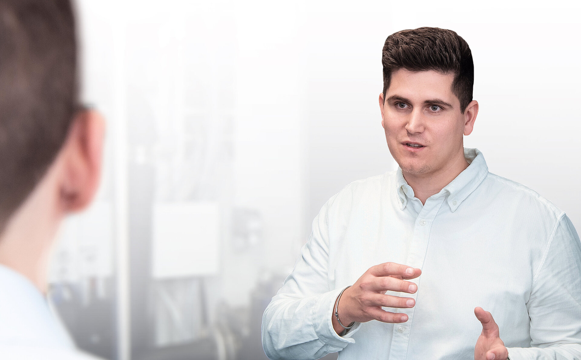 Zwei Personen in einem Gespräch, eine Person im Vordergrund und die andere im Hintergrund, beide in einem professionellen Umfeld.