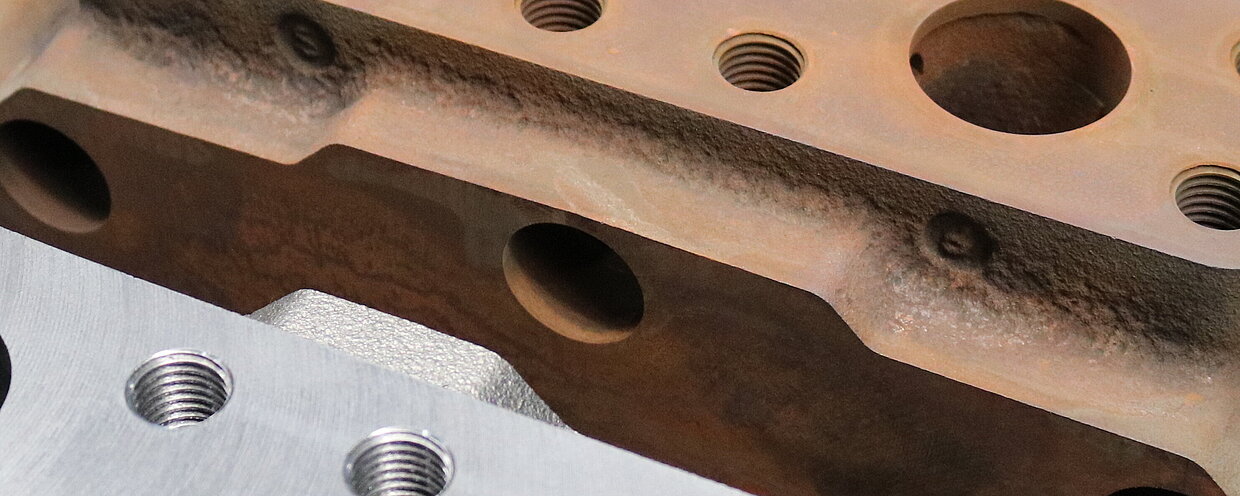 Drei Metallblöcke mit unterschiedlichen Oberflächenzuständen liegen nebeneinander auf einem Gitter. Der vordere Block ist glänzend und poliert, der mittlere ist rostig, und der hintere hat eine matte, unbehandelte Oberfläche. Die Blöcke haben mehrere Bohrungen und Gewinde.