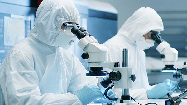 Zwei Personen in Reinraum-Schutzkleidung sitzen an einem Tisch und schauen durch Mirkroskope